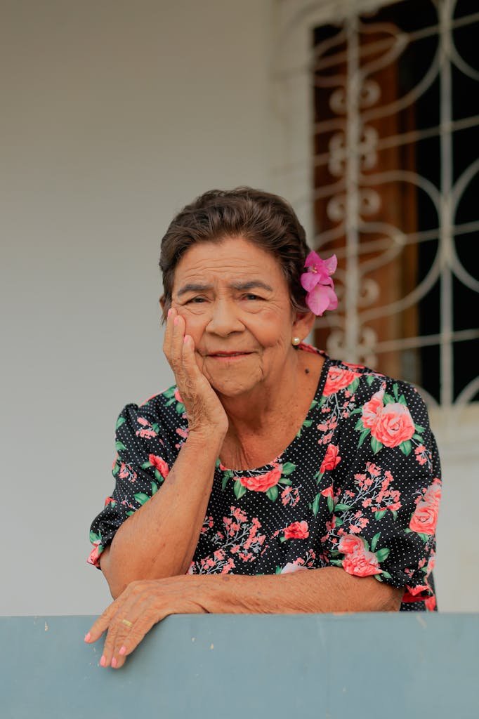 Charming portrait of an elderly woman with a floral dress and bright flower in hair, smiling gently outdoors.